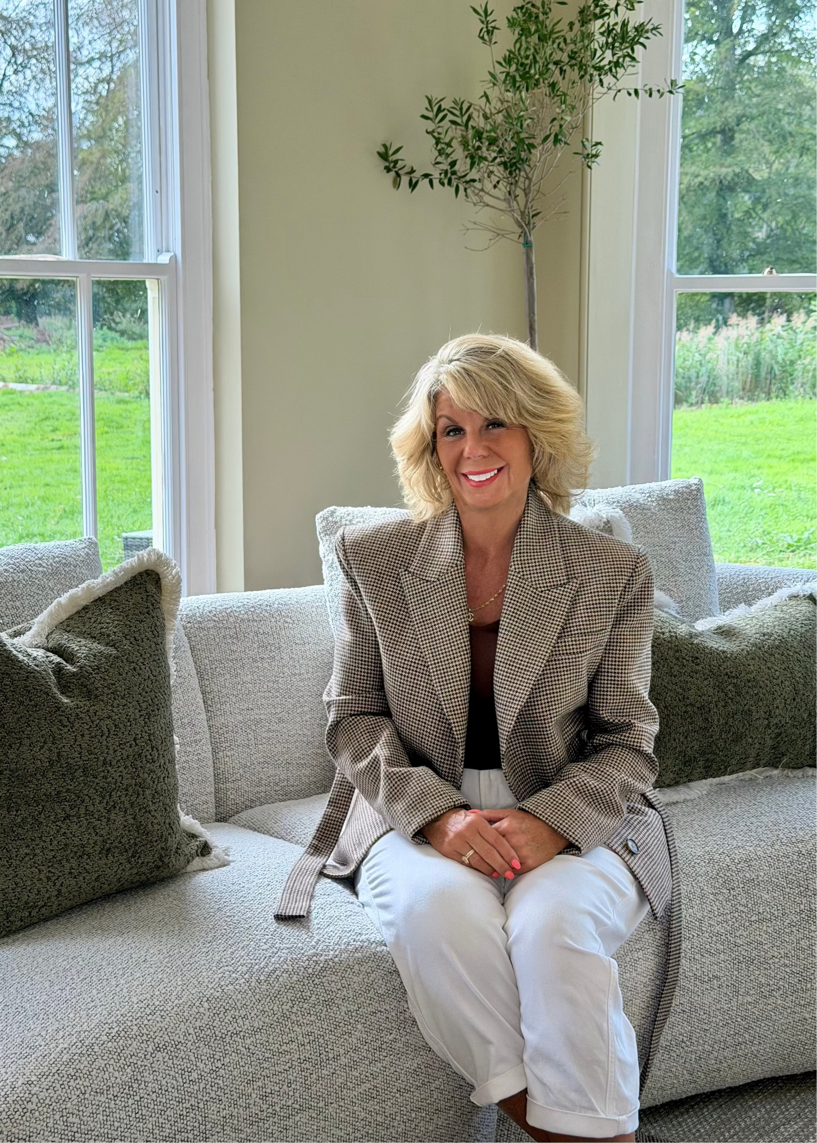 Woman seated on a sofa wearing the Brock houndstooth blazer with a black top and white trousers.
