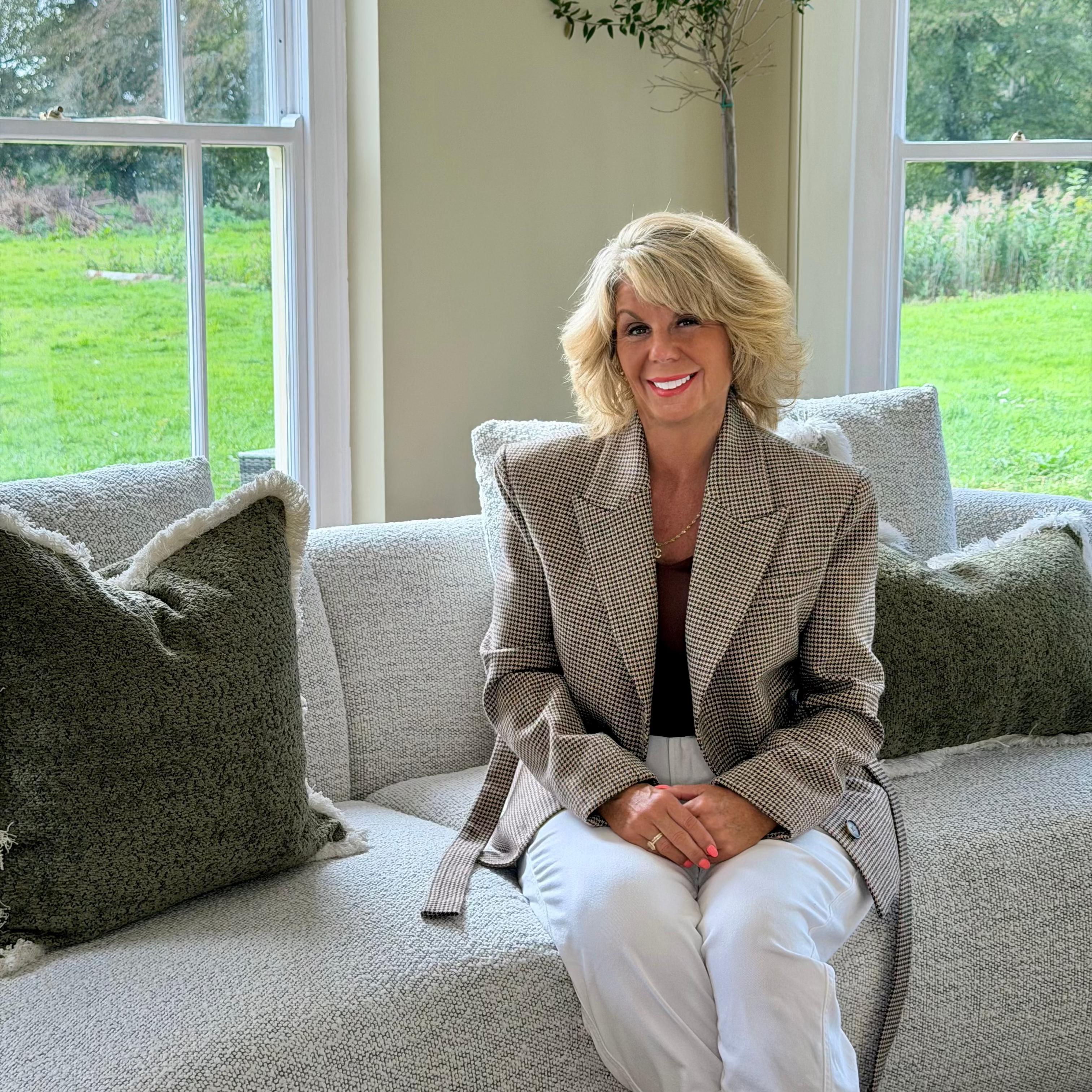 Woman seated on a sofa wearing the Brock houndstooth blazer with a black top and white trousers.